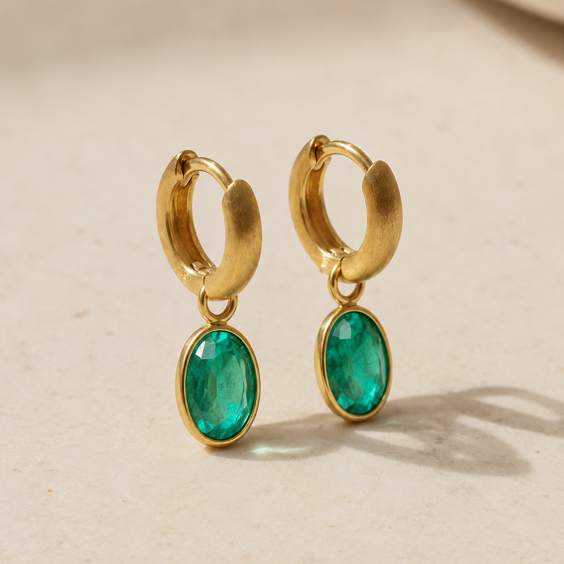 Gold hoop earrings with emerald green stones on a beige background